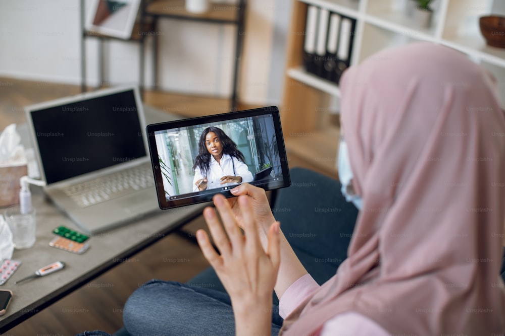 View from the shoulder of tablet screen with pleasant black woman doctor, consulting patient online. Young ill Muslim girl sitting on the couch at home, having an online consultation with doctor.