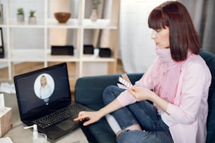 Jovem mulher fazendo chamada de vídeo para seu médico enquanto estava em casa. Close up do paciente com pílulas na mão, conferência com clínico geral no laptop. Menina doente na consulta on-line