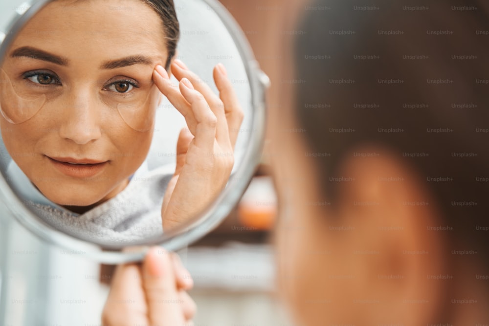 Konzentrierte attraktive junge Schönheitssalon-Kundin untersucht ihr Spiegelbild im kleinen runden Handspiegel