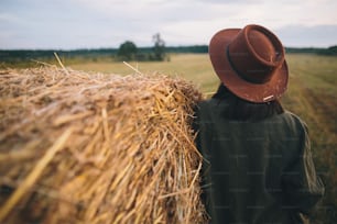 Stylish woman in hat with straw standing at haystack in summer evening field, back view. Atmospheric tranquil moment in countryside. Young female resting at hay bale.
