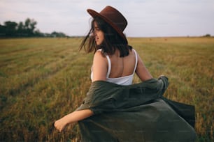 Beautiful carefree woman in hat walking and smiling in evening summer field. Young happy stylish female relaxing in countryside, enjoying evening. Atmospheric moment.
