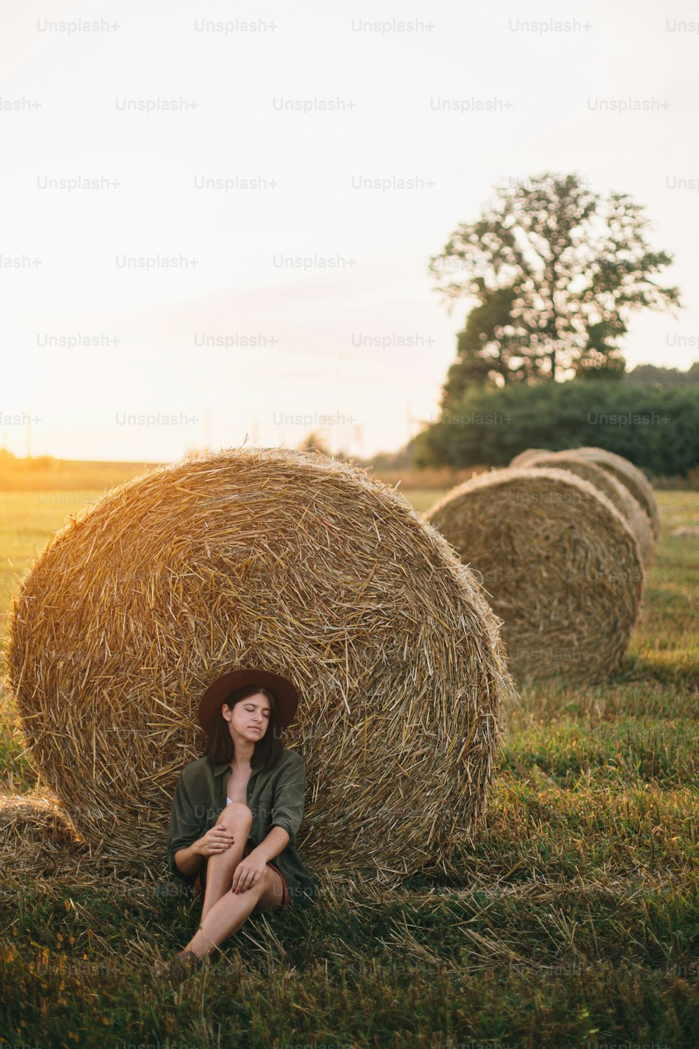 Beautiful stylish woman in hat sitting at haystacks enjoying evening in sunny summer field. Atmospheric tranquil moment in countryside. Young female relaxing at hay bale in warm sunset