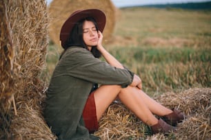 Beautiful stylish woman with herb in mouth and in hat sitting at haystack in summer evening field. Sexy young female relaxing at hay bale, summer vacation in countryside.