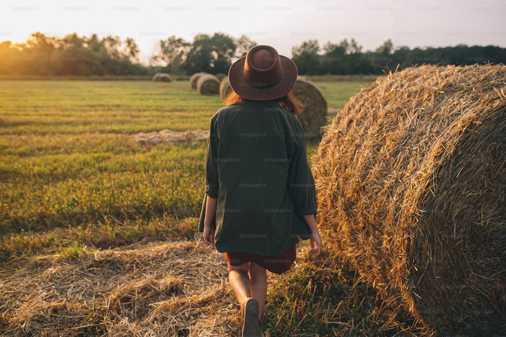 夏の野原の夕暮れの光の中で干し草の山を歩く帽子をかぶった美しいスタイリッシュな女性。田舎の雰囲気の静かなひととき。暖かい日差しの中で干し草の俵で夜を楽しむ若い女性