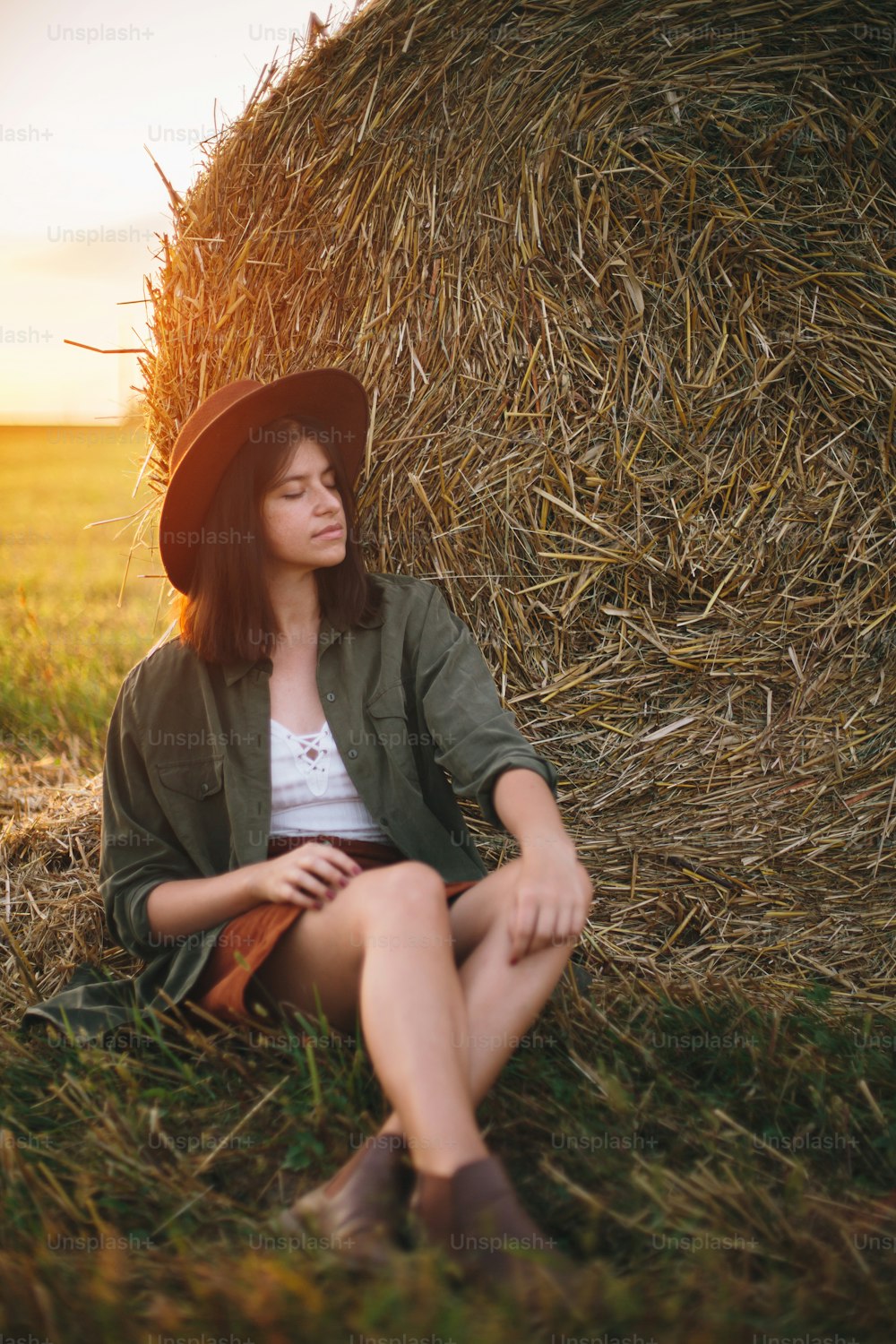 Hermosa mujer elegante con sombrero sentada en pajares disfrutando de la noche en el campo soleado del verano. Momento de tranquilidad atmosférica en el campo. Hembra joven relajándose en la paca de heno en la puesta del sol cálida