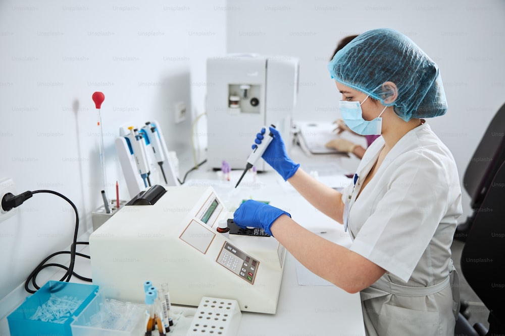 Seitenansicht eines Biochemikers in einer Maske mit einer automatisierten Pipette in einer Hand am Labortisch sitzend