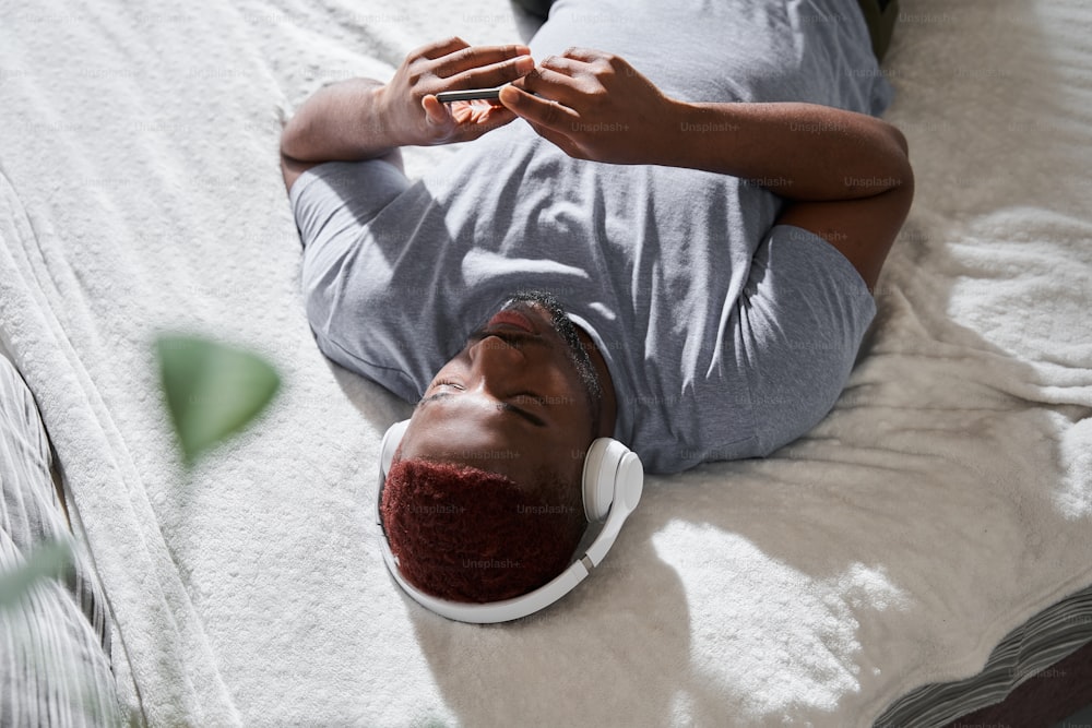 Close up view of the multiracial man wearing headphones laying at the bed and listening music while switches songs at his smartphone. Man spending his calm great morning. Stock photo