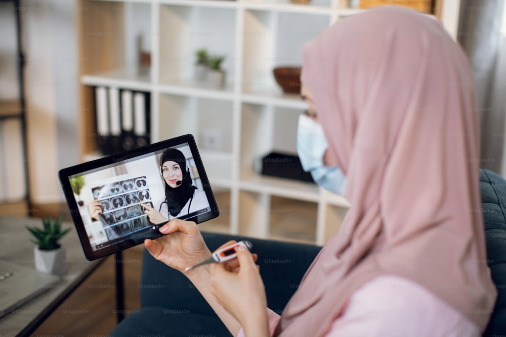 Side blurred view of sick young Muslim woman holding thermometer in hands, doing a medical consultation online with female Arabian gp doctor on digital tablet at home