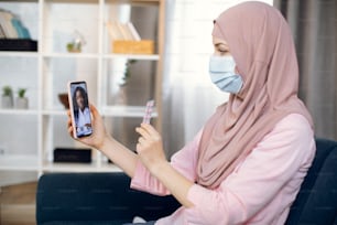 Mulher muçulmana doente de hijab e máscara de proteção, segurando celular em uma mão e pílulas em outra. Mulher terapeuta médica africana em chamada de videoconferência on-line, ajuda sua paciente do sexo feminino.