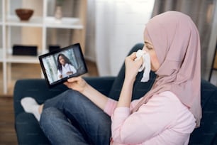 Sick Muslim woman patient suffering from cold, covid and running nose, sitting on sofa at home, talking with her doctor, young confident African American woman, using tablet pc
