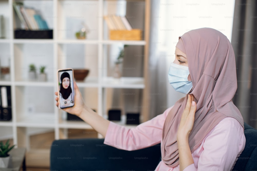 Muslim young woman patient with sore throat, having video chat with Arabian female doctor via smartphone app, sitting on sofa at home. Tele medicine, technology and healthcare concept.