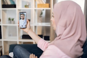 Muslimische junge Frau, die im Hijab leidet, Video-Chat mit männlichem Arzt über Smartphone-App, zu Hause auf dem Sofa sitzend. Telemedizin, Technologie und Gesundheitskonzept.