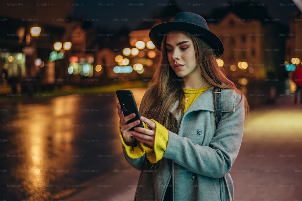 Mujer hermosa joven usando un teléfono inteligente y siendo iluminada por la luz de la pantalla del dispositivo mientras camina por la ciudad por la noche