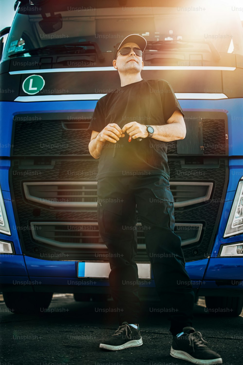 모자와 선글라스를 쓴 전문 트럭 운전사가 크고 현대적인 트럭 함대 앞에 자신 있게 서 있습니다. 밝고 화창한 날. 사람과 교통 개념.