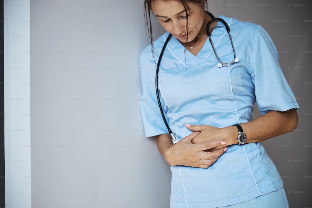 Nice young woman doctor leaning on the wall and placing hands on stomach while suffering from abdominal pain at work