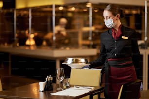 Foto dello spazio di copia di una cameriera professionista che utilizza una maschera protettiva mentre serve il cibo ai tavoli del ristorante