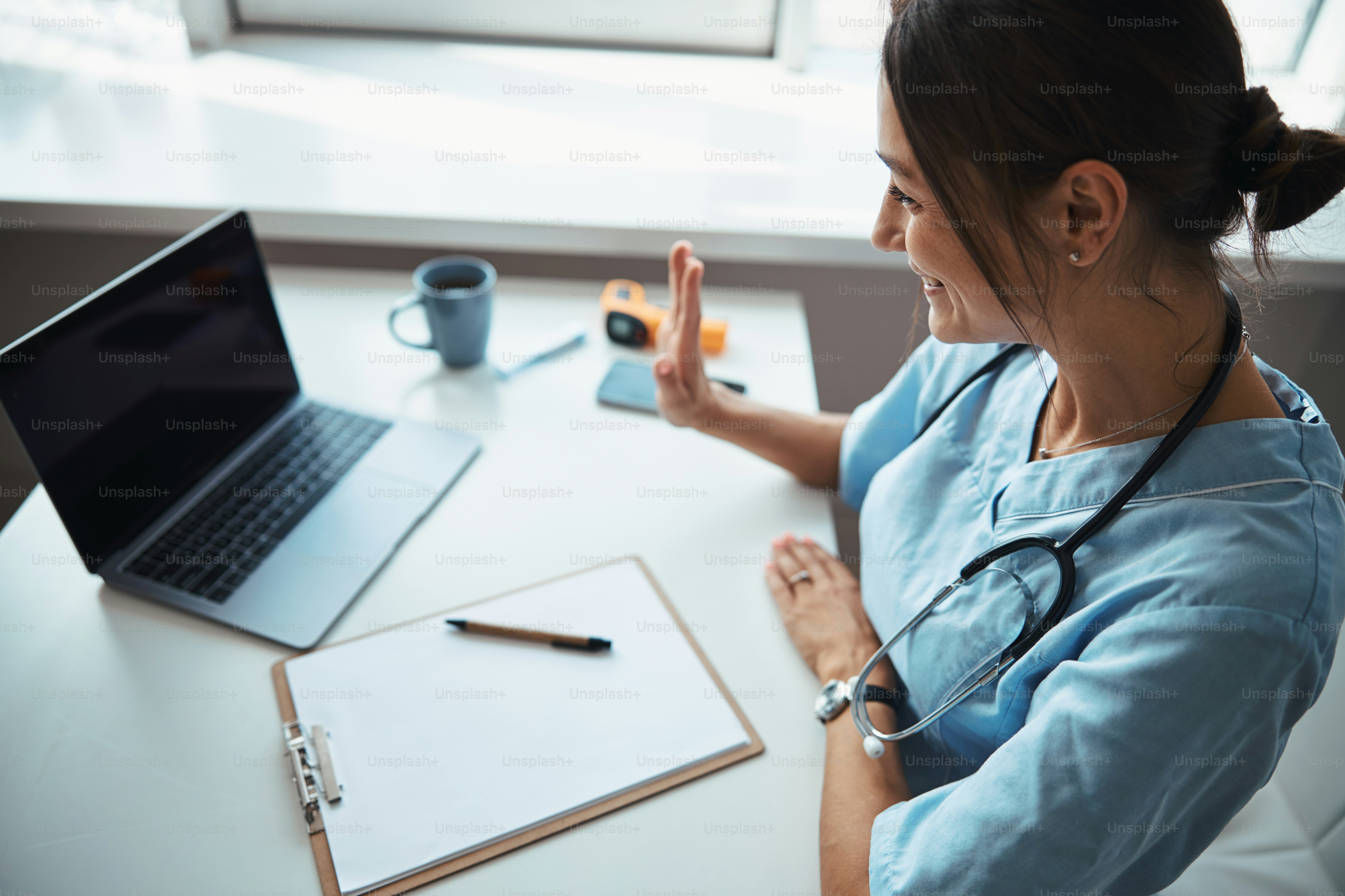 Joyful young woman physician doing hello gesture and smiling while sitting at the table with notebook and giving online consultation