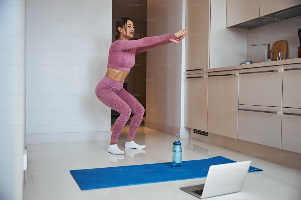 Beautiful sporty lady doing squats and smiling while having morning workout in her apartment