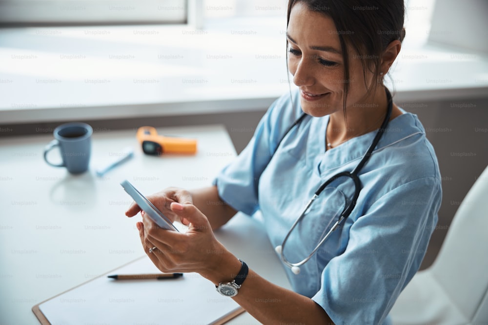 Charming young woman physician reading message on smartphone and smiling while sitting at the table with clipboard
