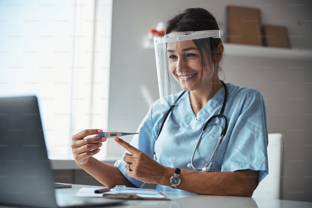 Cheerful young woman physician wearing protective face shield mask while holding digital thermometer and talking with patient online during pandemic