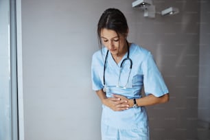 Bonita trabajadora médica joven de pie junto a la pared y colocando las manos en el estómago mientras tiene dolor de estómago en la clínica