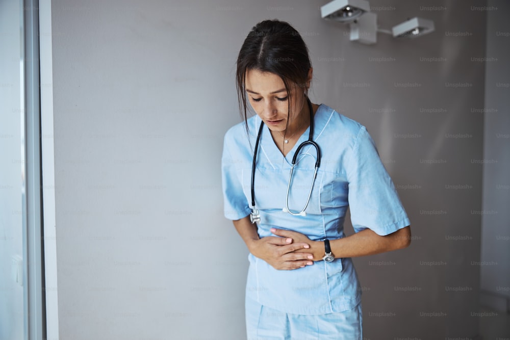 Nice young woman medical worker standing by the wall and placing hands on stomach while having stomachache in clinic