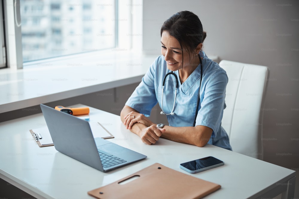 Charming young woman physician sitting at the table with notebook and smiling while giving online consultation in clinic