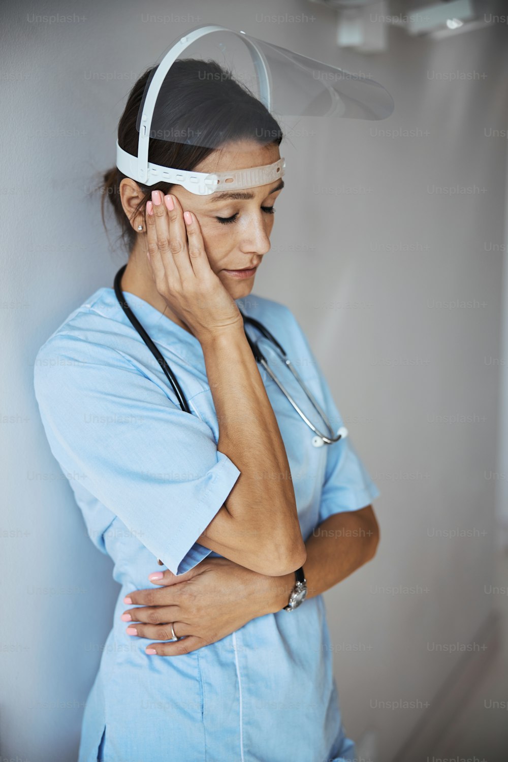 Nette junge Ärztin mit Schutzmaske und medizinischer Uniform, während sie an der Wand lehnt und unter Kopfschmerzen leidet