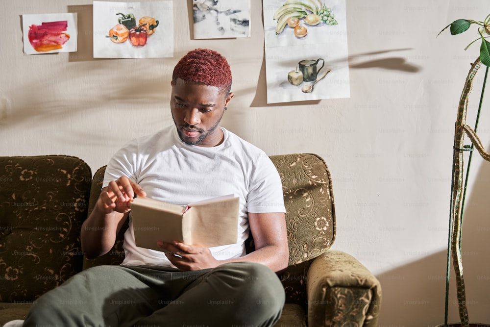 Waist up portrait view of the multiracial man relaxing on sofa at home while reading his favorite book. Guy involved at the novel at the couch. Stock photo