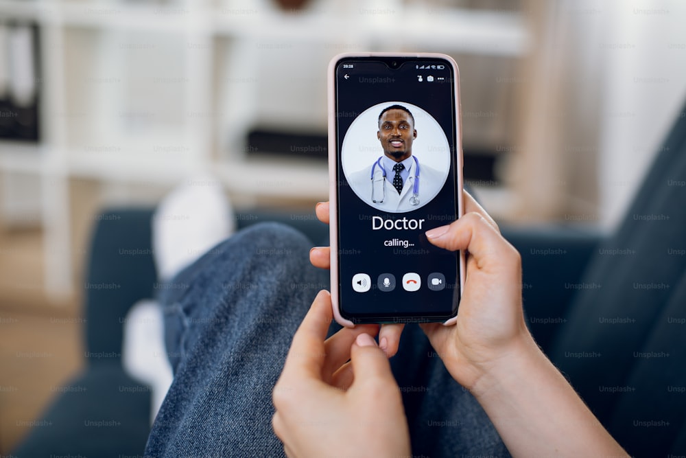 Hands of female patient, sitting on blue couch at home, holding smartphone calling to male African American doctor for online video chat. Medical consultation online.