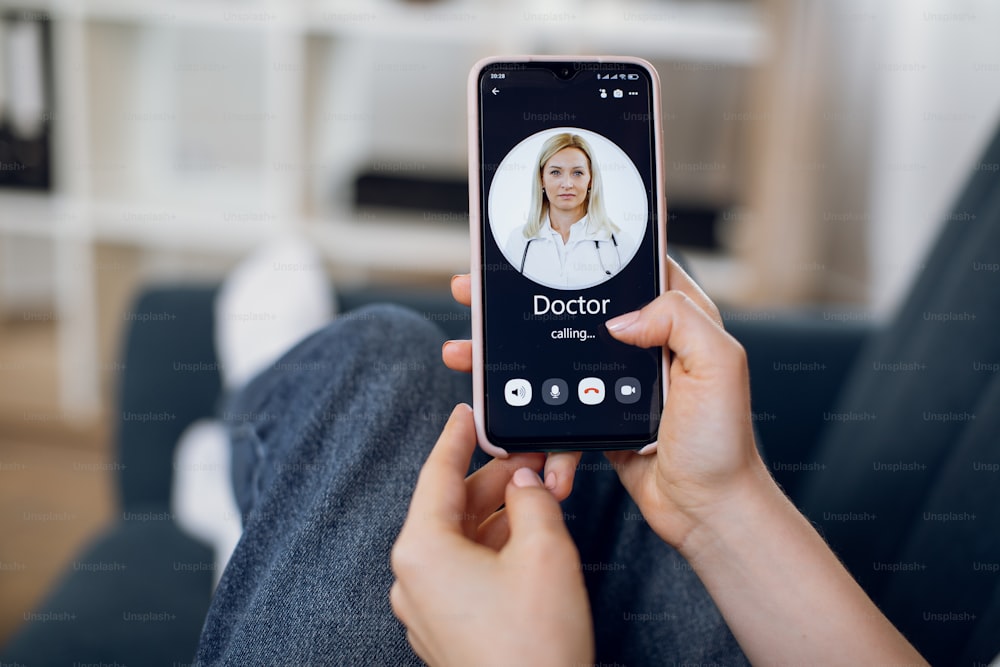Virtual medical consultation. Unrecognizable woman, sitting on couch at home, calling to doctor via video call on cellphone. Close up of smartphone screen with picture of blond mature female doctor