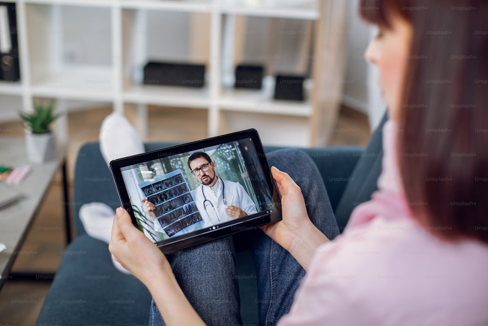 Telemedicine. Remote medicine concept. Back shoulder view of tablet pc screen with confident male doctor, holding xray scan and consulting his patient, staying at home. Woman sits on sofa at home.