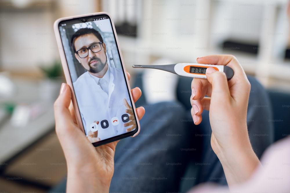Online medicine concept. Close up screen view of confident bearded doctor communicates with his patient via a video call on a smartphone. Sick woman with thermometer takes gp remote consultation.