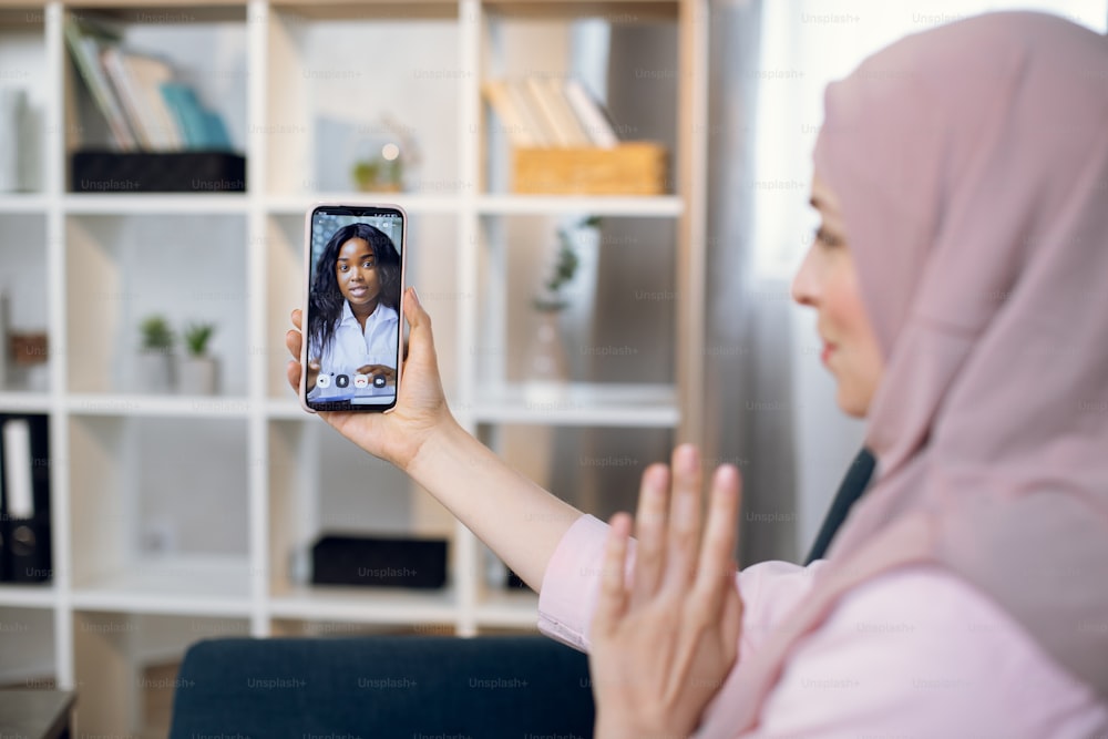 Telemedicine, online appointment concept. Side view of pretty young Muslim woman, wearing hijab, sitting on sofa at home, having video chat with female afro doctor by phone.