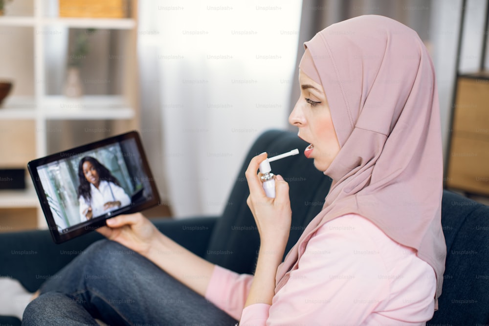 Young sick Muslim woman sits on couch at home and holds a spray for sore throat. Blur view of African female doctor talking to patient by video call on tablet. Online quarantine medical consultation.