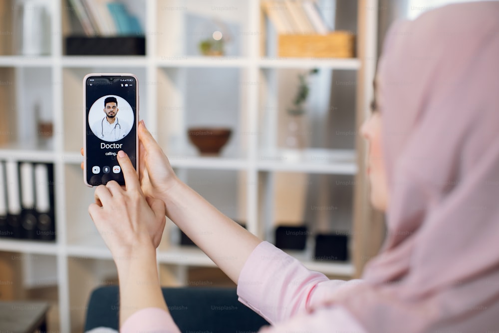 Close up blurred back view of young Muslim woman sitting at home, calling for online consultation with male Indian Arabian doctor on cell phone. Telemedicine and healthcare concept.