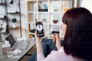 Concept of telemedicine and patient consulting online. Sick woman feeling bad at home, measuring body temperature, calling to male indian doctor using smartphon. Focus on phone screen