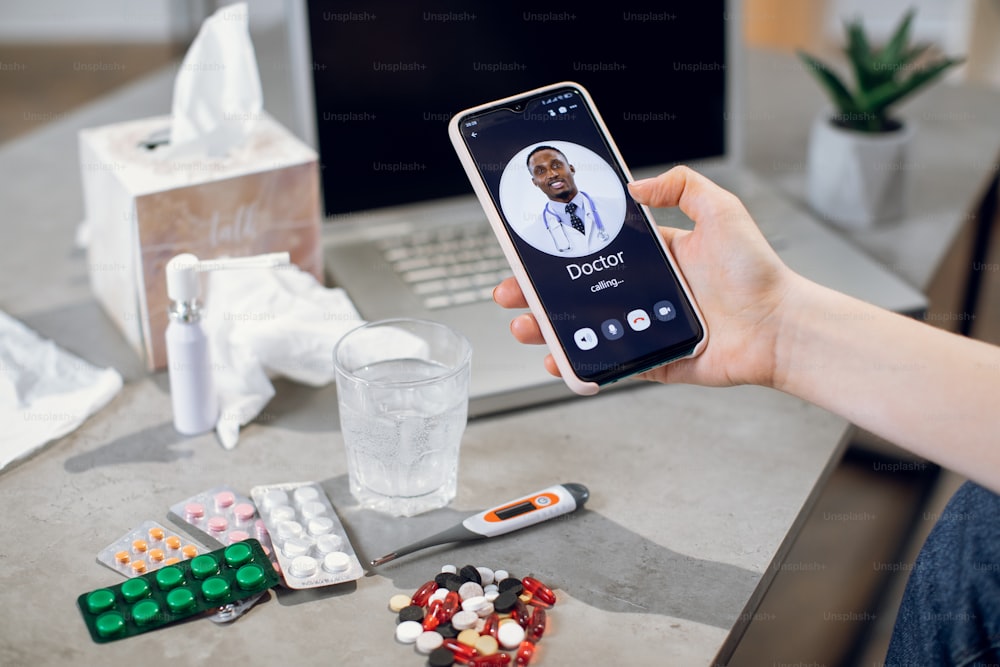 Hands of young unrecognizable patient, holding smartphone and calling to male afro american general practitioner for online consultation. Table with pills, thermometer and water.