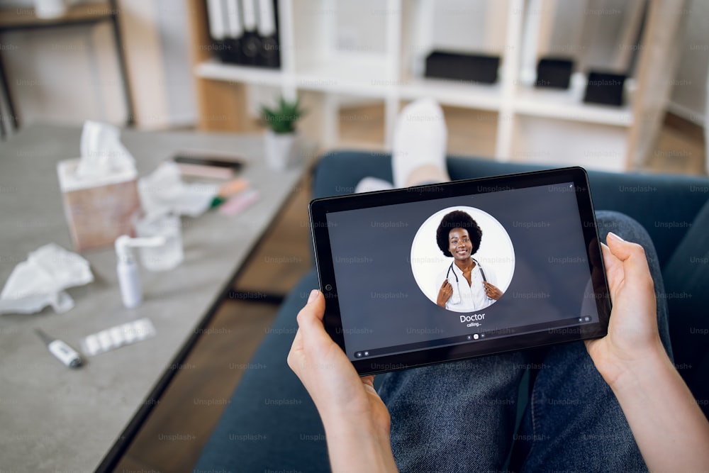 Tablet screen view, close up. Top view of unrecognizable female patient, sitting with digital tablet pc on couch and making video call to young confident black medical practitioner from home.