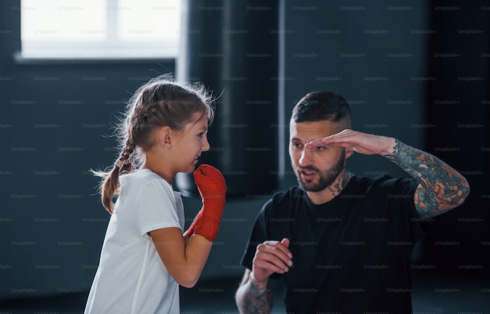 Basic punches. Young tattooed boxing coach teaches cute little girl in the gym.