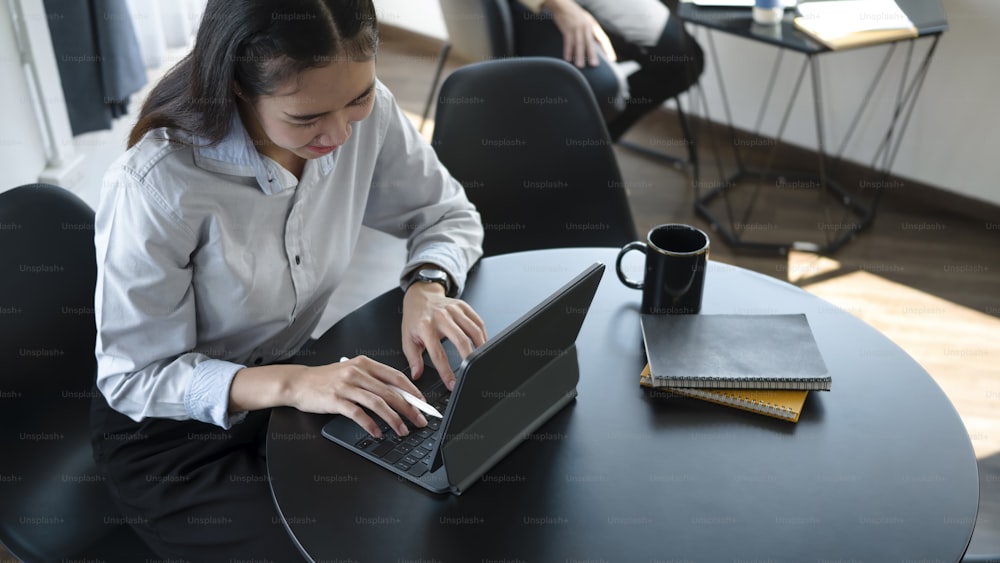 Lächelnde junge asiatische Angestellte, die im Büroraum sitzt und ein Computer-Tablet benutzt.