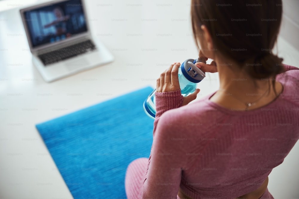 Close up of female person sitting on yoga mat and holding bottle of water while watching training on laptop