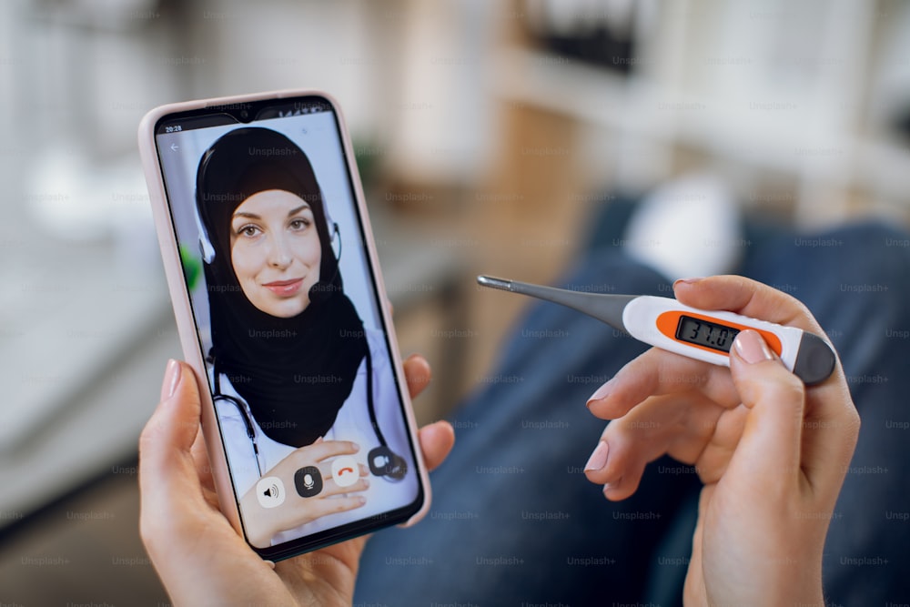 Telemedicine concept, app for online medical service. Close-up of a female Muslim doctor on phone display during consultation with woman patient, holding thermometer measuring body temperature.