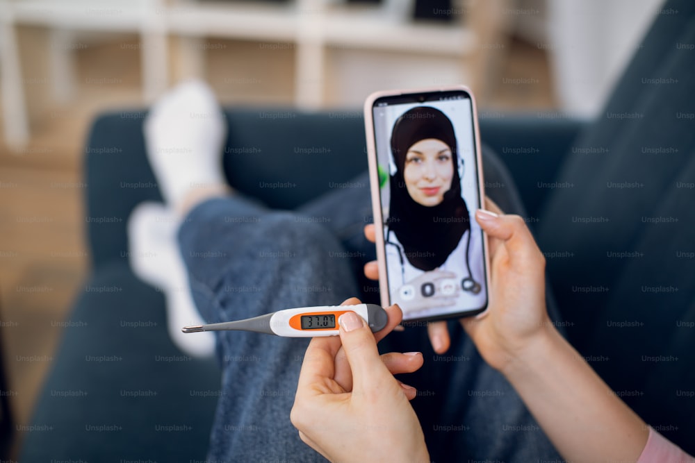 Unrecognizable sick female patient, sitting on sofa at home with thermometer in hand, videochatting with muslim doctor in hijab on mobile phone. Self control of treatment at home.