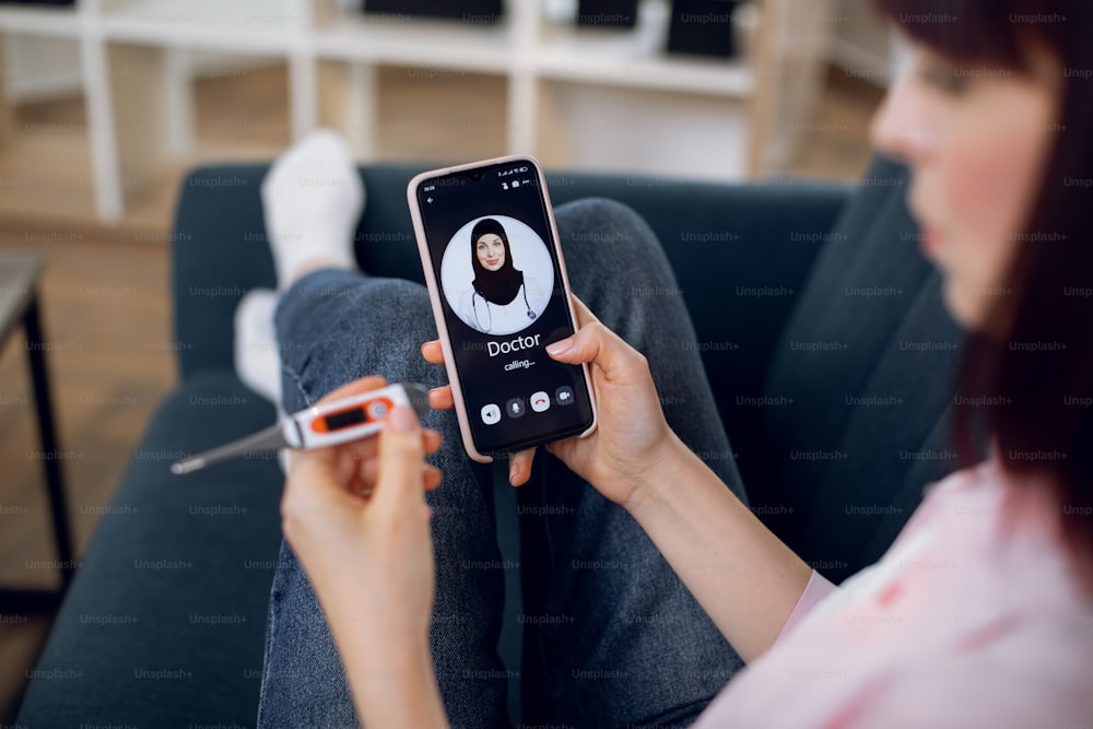 Sick young woman feeling bad and having fever, holding thermometer, making video call to muslim female doctor, using phone at home. Telemedicine and patient counseling online concept.