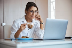 Beautiful woman holding cup of hot drink and smiling while sitting at the table with modern notebook in office