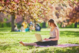 Hermosa joven trabajando en su computadora portátil en el parque durante la temporada de floración de los cerezos. Mujer joven en el famoso Parque de Sceaux cerca de París, Francia. Concepto de trabajo independiente, a distancia o a distancia