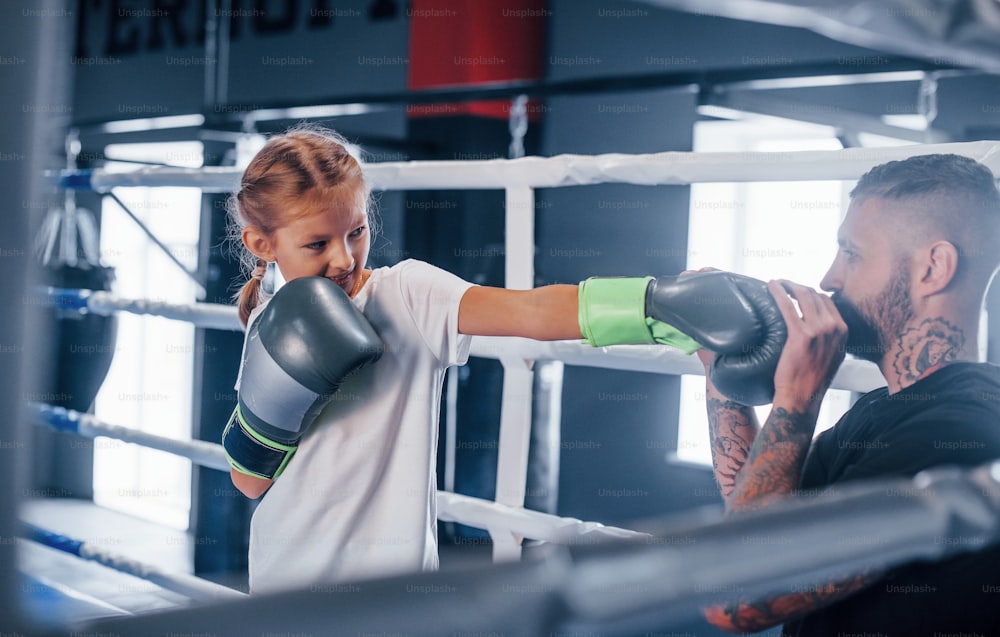 Sparring miteinander auf dem Boxring. Junger tätowierter Boxtrainer unterrichtet süßes kleines Mädchen im Fitnessstudio.