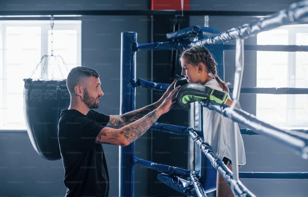 Konzeption von Hilfestellungen. Junger tätowierter Boxtrainer unterrichtet süßes kleines Mädchen im Fitnessstudio.