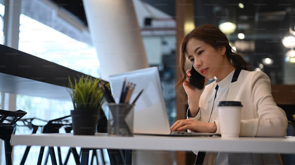 Asian woman entrepreneur talking on mobile phone and using computer laptop at modern workplace.